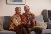 An older couple sitting on the couch. Both smiling and looking at a tablet. She has her hand on his shoulder.